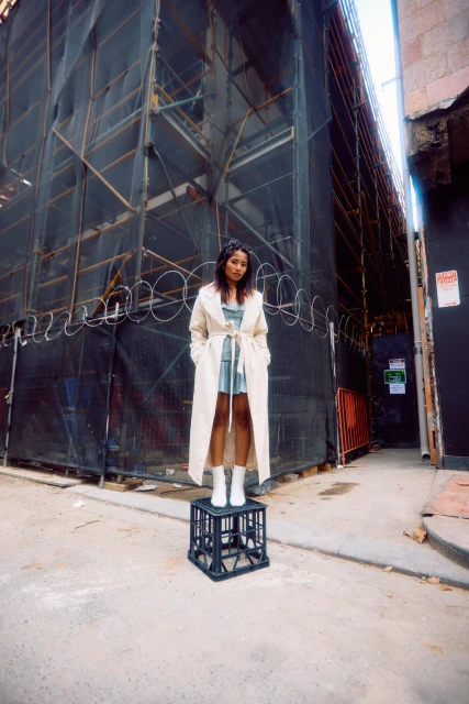 A young woman stands on a black plastic crate in an urban alleyway, wearing a long trench coat and white boots, posing for a fashion photograph.
